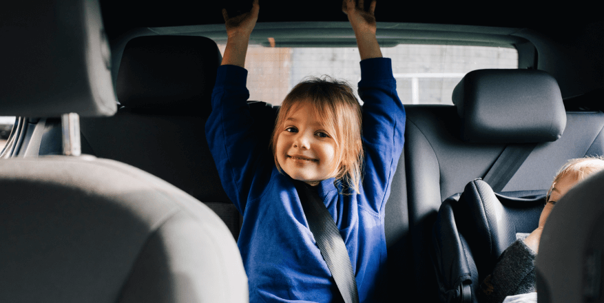 A child sits in the back seat of a car with arms raised, smiling.